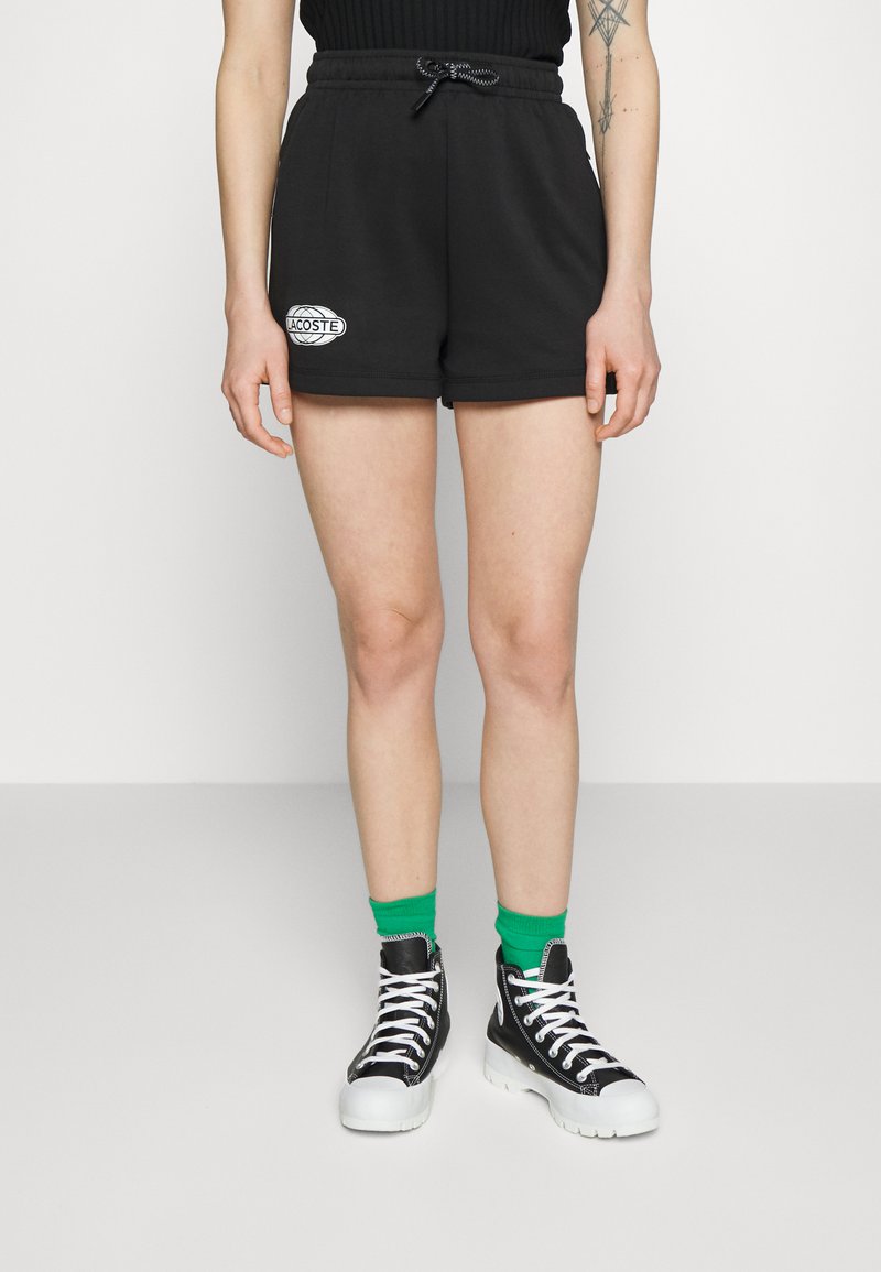 Calções de algodão pretos com uma cintura elástica e cordão, apresentando um patch do logótipo branco da Lacoste. Combinados com ténis pretos de cano alto e meias verdes.