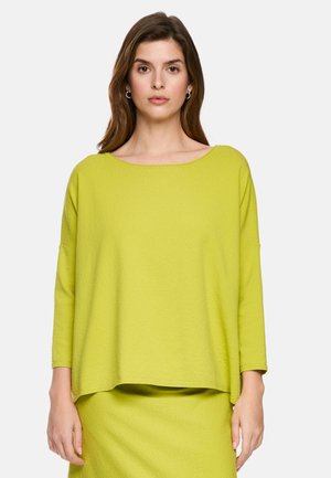 Woman with long brown hair wearing hoop earrings and a loose-fitting lime green long-sleeve top and matching skirt, facing forward.