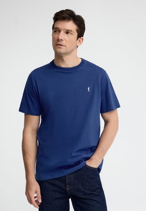 Uomo con capelli scuri che indossa una maglietta blu a maniche corte con un piccolo logo bianco da golf, in piedi con una mano in tasca, guarda verso sinistra.