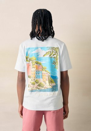 Man with shoulder-length twists wears white t-shirt with colorful coastal village print and pink shorts, standing against pale background.