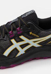 Baskets noires de sport avec une tige en maille grise, accents jaunes et bleu clair, boucles de lacet violettes, et semelle noire texturée présentant un motif de traction.