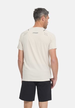 Mann trägt hellbeiges kurzärmeliges Hemd und schwarze Shorts, steht mit dem Rücken zu einem schlichten weißen Hintergrund.