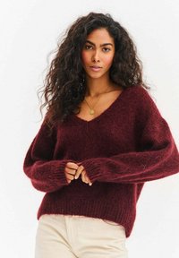 Bordowy, oversize sweter wykonany z miękkiej, teksturowanej tkaniny, z głębokim dekoltem w serek i szerokimi rękawami, noszony na jasnych spodniach.