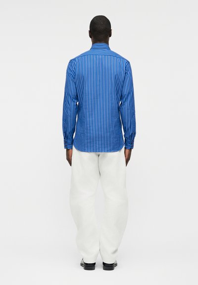 Άνδρας που στέκεται γυρισμένος πλάτη, φορώντας μπλε πουκάμισο με κάθετες ρίγες μακριάς μανίκας, άσπρο παντελόνι και μαύρα παπούτσια, μπροστά σε απλό φόντο.