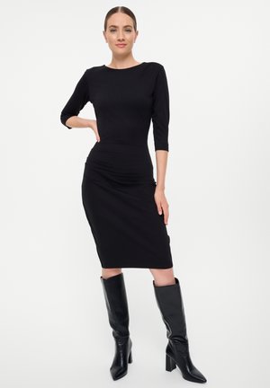 Robe noire ajustée avec des manches trois-quarts, longueurs jusqu'aux genoux et détails froncés. Portée avec des bottes noires à talons jusqu'aux genoux.