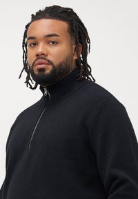 Mann med dreadlocks og skjegg iført en svart zip-up genser med ribbet krage, ser fremover mot en ensfarget hvit bakgrunn.