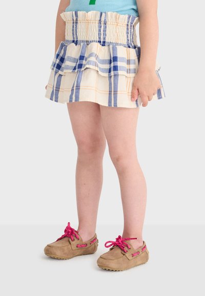Enfant portant une jupe à volants à carreaux bleus et blancs avec des mocassins marron clair ornés de lacets rose vif, debout sur un fond uni.