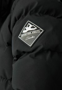 Svart pufferjacka med kviltad textur. Har en silverfärgad logotyp som visar berg och texten: "Äventyret väntar Clarks."
