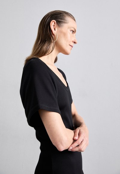 Schwarzes, geripptes Kleid mit V-Ausschnitt und kurzen Ärmeln. Das Model steht seitlich, die Arme verschränkt. Einfaches und figurbetontes Design mit glatter Textur.
