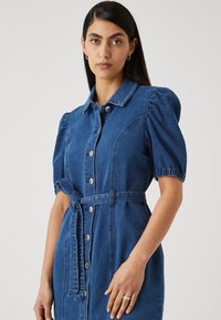 Μάξι φόρεμα από τζιν με γιακά πουκαμίσου, κουμπιά μπροστά, κοντά φουσκωτά μανίκια και ζώνη στη μέση. Σκούρο μπλε χρώμα με λεία υφή.