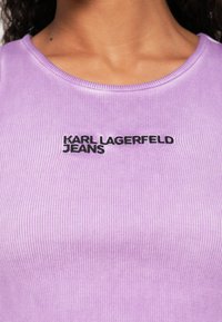 Záber na hornú časť tela osoby oblečenej v fialovom rebrovanom tričku s výšivkou "KARL LAGERFELD JEANS" čiernou farbou uprostred na hrudi.