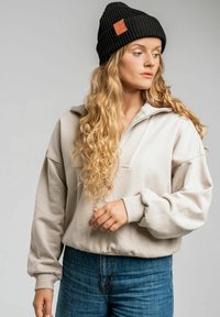 Beżowy pulower z kołnierzem na pół-zamek, z mankietami i prążkowaną teksturą, połączony z czarną prążkowaną czapką beanie z skórzaną metką.