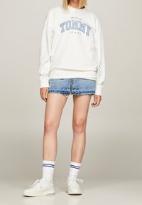 Žena má na sobě bílou mikinu "Tommy Jeans", světle modrou džínovou mini sukni s předním zipem, bílé ponožky s modrými pruhy a bílé tenisky.