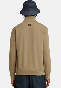 Felpa beige con colletto alto, polsini e orlo a costine. Presenta un piccolo etichetta con logo sul retro, indossata con un cappello a secchiello di denim.