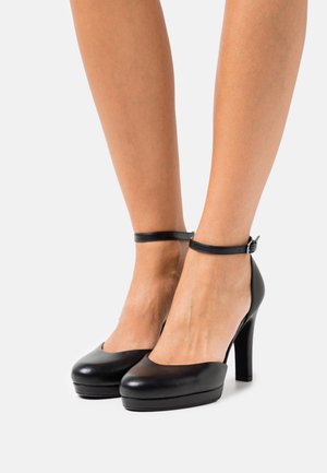 Chaussures à talons hauts en cuir noir avec un bout arrondi, une bride de cheville avec une boucle et une semelle compensée, affichant une texture lisse et brillante.