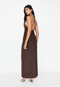 Braunes, ärmelloses Maxikleid mit niedrigem Rückenausschnitt, Raffungen an der Taille und glatter Textur. Kombiniert mit offenen, hohen Sandalen.