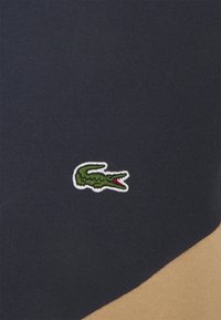 Zelené logo krokodýla vyšívané na pozadí látky s námořnickou a béžovou barevnou blokací.