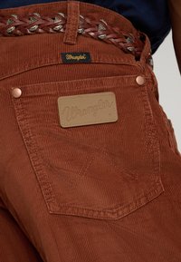 Rudi korduroviniai kelnės su odiniu Wrangler ženkliuku, austu odiniu diržu ir kontrastingomis siūlėmis ant galinės kišenės.