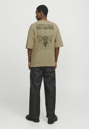 Camiseta oversize color caqui con un gráfico de una polilla y el texto "OBRA DE ARTE CONTEMPORÁNEA" en la parte trasera; combinada con pantalones de mezclilla oscuros.