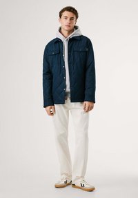 Chaqueta acolchada navy con dos bolsillos en el pecho, superpuesta sobre una sudadera ligera gris claro. Combinada con pantalones blancos y zapatillas blancas con detalles en negro.