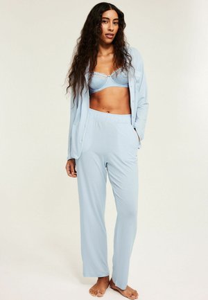 Kvinde med langt krøllet hår iført lyseblå pyjamas og en matchende bh, stående barfodet mod en ensfarvet baggrund.