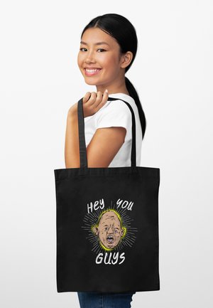 Donna sorridente con capelli neri raccolti tiene una borsa tote nera con un volto da cartone animato e il testo "Hey you guys" su sfondo bianco.
