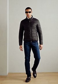 Doudoune noire avec fermeture éclair, associée à un jean bleu foncé et des chaussures noires. Modèle portant des lunettes de soleil, se tenant dans un cadre intérieur minimaliste.