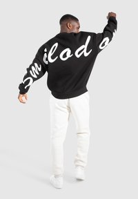 Felpa nera con testo bianco in corsivo sulla schiena, abbinata a pantaloni della tuta bianchi e sneakers bianche. Vestibilità ampia con orlo e polsini a costine.