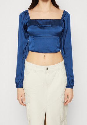 Royal blue satiini crop-top, jossa on neliön muotoinen pääntie, pitkät puffihihat ja kerrostettu yksityiskohta. Yhdistetty vaaleanväriseen hameeseen.