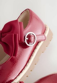 Scarpa in pelle verniciata rossa con finitura lucida, dotata di un grande fiocco a coste e una fibbia d'argento sulla cinghia. Suola in tessuto beige chiaro.