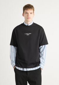 Crna majica s okruglim izrezom s logom "Calvin Klein JEANS" u bijeloj boji, nosi se preko majice na plavo pruge. Glatka pamučna tkanina, ležeran kroj.