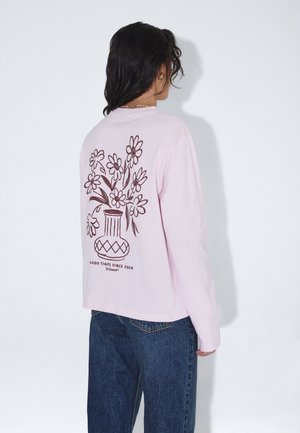 Persona con cabello oscuro lleva una camisa de manga larga rosa claro con un diseño de jarrón floral marrón y el texto "Good Times Since 2004" en la espalda, combinada con jeans azules.