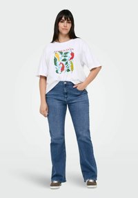 T-shirt graphique blanche avec un design de poivron coloré et le texte "PIMENTÓN", associée à un jean évasé bleu et des baskets marron.