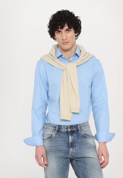 Lichtblauw button-up shirt met opgerolde mouwen, gecombineerd met een crèmekleurige trui die om de schouders is gedrapeerd, en versleten blauwe jeans.