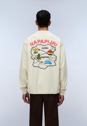 Hombre de espaldas que lleva una camisa de manga larga color crema con diseño de mapa colorido y texto rojo "NAPAPIJRI GLOBAL MAPPING" en la espalda.