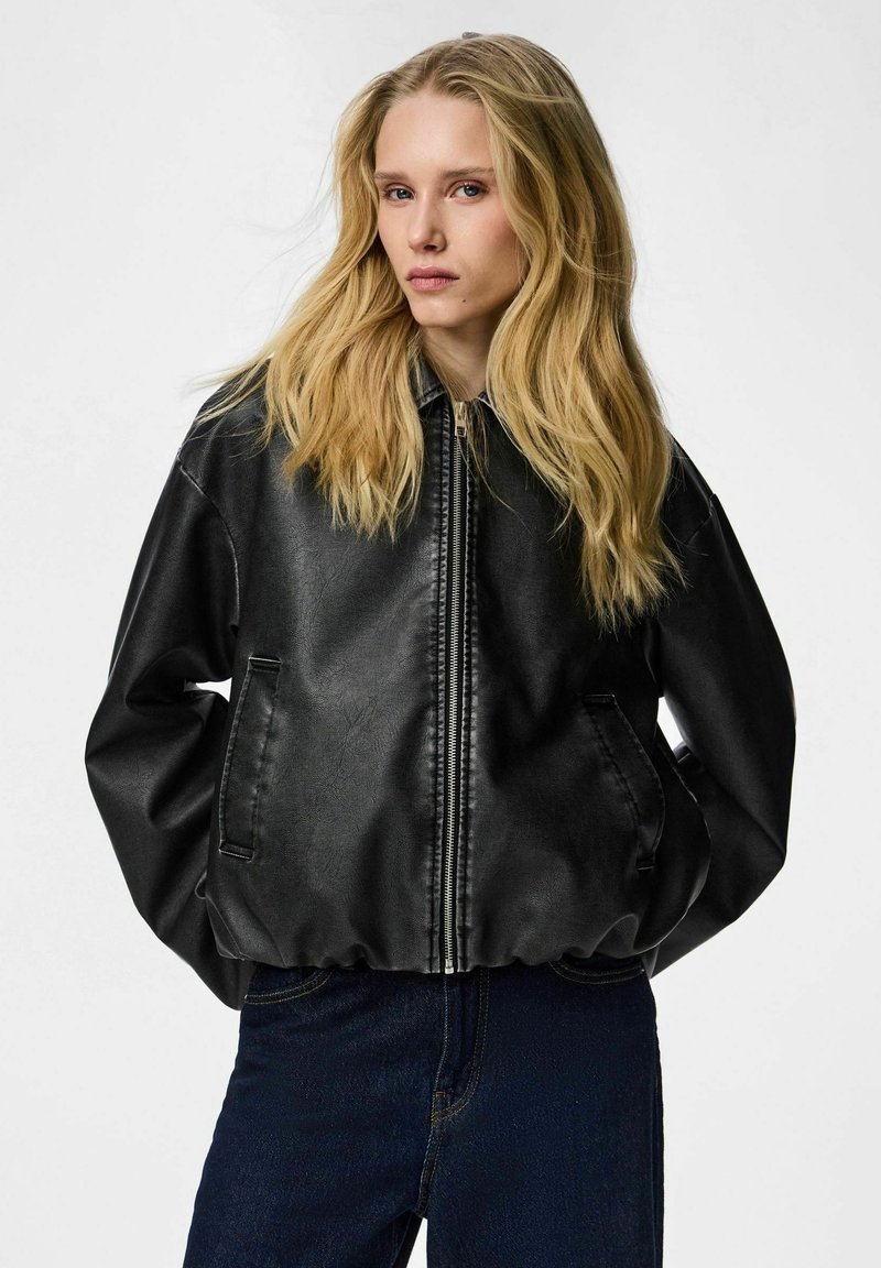 Femme blonde aux cheveux longs portant une veste en cuir noire zippée et un jean bleu foncé, sur un fond blanc uni.