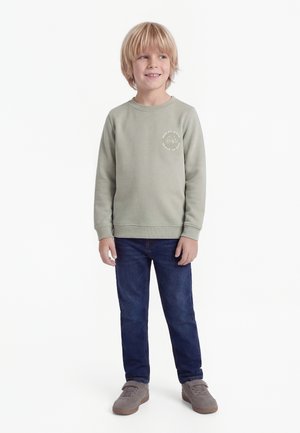 Ragazzo biondo che indossa una felpa verde chiaro con grafica, jeans blu scuro e sneakers grigie, in piedi e sorridente leggermente di lato.