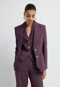 Bordeauxkleurige op maat gemaakte blazer met brede kleppen, gouden knopen en zijzakken, met daaronder een bijpassende gelaagde jas. Zachte textuur.