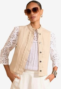 Beige sleeveless vest with scalloped edges and silver buttons, worn over a white eyelet shirt with puffed sleeves and detailed patterns.