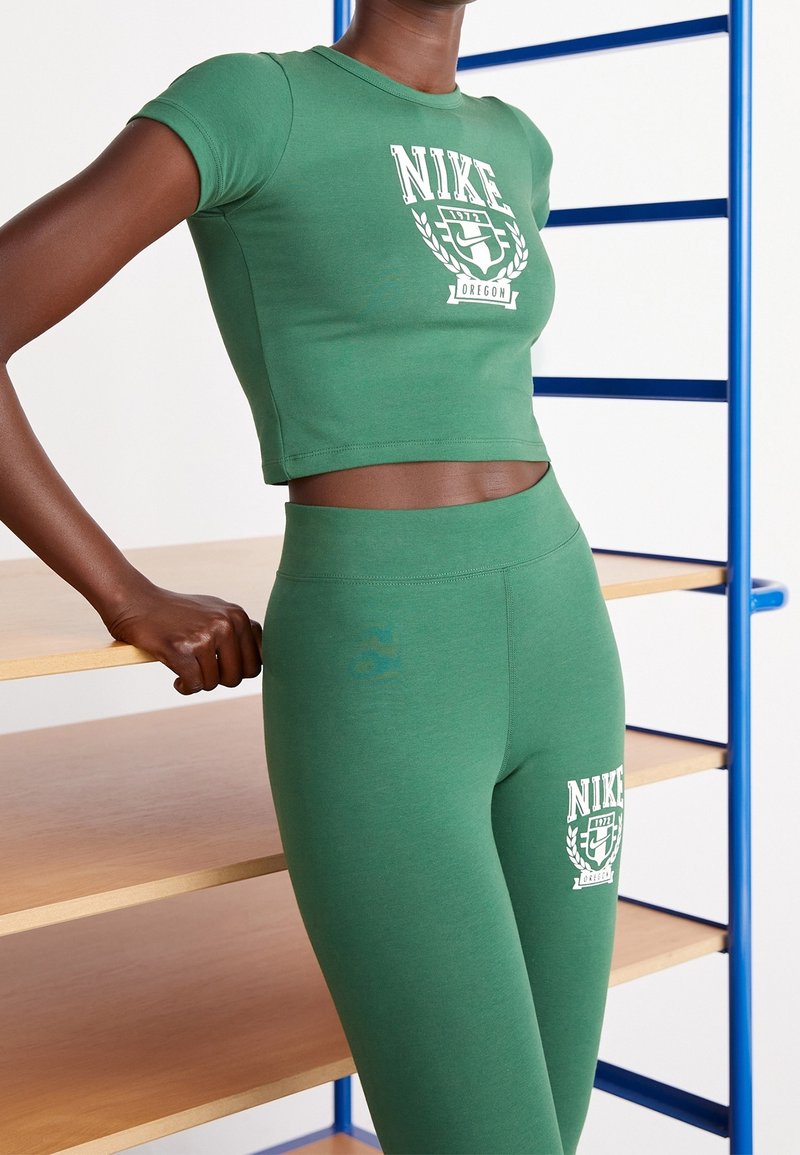 Osoba v zeleném crop topu Nike a odpovídajících legínách se opírá o dřevěné regály s modrým kovovým rámem.