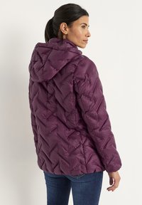 Kvinna i en lila vadderad jacka med huva och bl�å jeans, vänd sideways och tittar över axeln mot en enkel bakgrund.