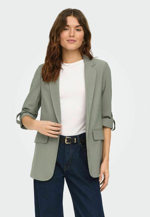 Donna con capelli castano ondulati indossa un blazer grigio chiaro sopra una camicia bianca, jeans blu scuro a vita alta e una cintura nera, posando con la mano vicino al blazer.
