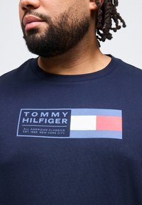 Κοντινό πλάνο ενός ατόμου που φοράει μπλε σκούρο πουκάμισο με το λογότυπο Tommy Hilfiger και κείμενο που γράφει "All American Classics Est. 1985 New York City."