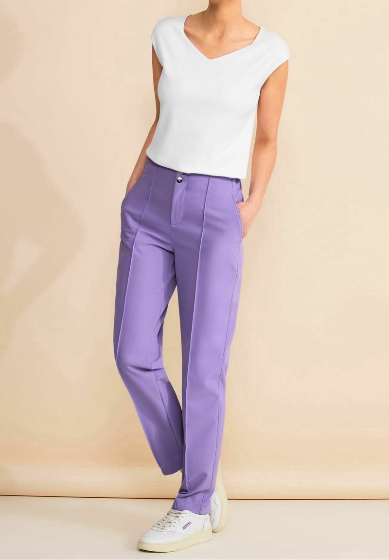 Nainen, jolla on päällään valkoinen V-kaula-aukkoinen hihaton paita ja korkeavyötäröiset laventelinväriset housut, valkoiset lenkkarit jalassa, seisoo kädet taskuissa vaaleanbeigeä taustaa vasten.