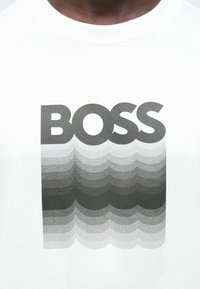 Bela srajca z izrazitim črnim napisom "BOSS" nad bledenim gradientnim vzorcem, ki spominja na valove v odtenkih sive.