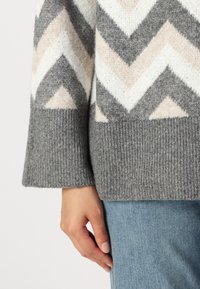 Grå genser med chevron-mønster og kremfargede og lyserosa detaljer. Ribbede mansjetter og hem med myk, teksturert stoff. En hånd synlig på nederste kant.