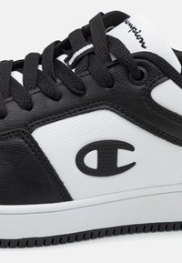 Sort og hvid sneaker lavet af læder og syntetiske materialer, med runde sorte snørebånd, logo på siden og teksturerede hvide paneler.