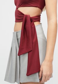 Tumedat punast värvi lühike t-särk, millel on vöökohal sõlmitud detail, koos helehalli volditud seelikuga. Kootud kangas, sileda tekstuuriga.