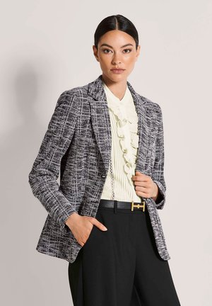 Vrouw met glad haar draagt een zwart-wit gestructureerde blazer over een gerimpelde crèmekleurige blouse, gecombineerd met een zwarte broek en een zwarte riem.