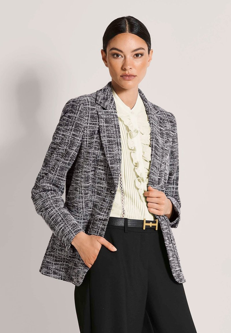 Vrouw met glad haar draagt een zwart-wit gestructureerde blazer over een gerimpelde crèmekleurige blouse, gecombineerd met een zwarte broek en een zwarte riem.